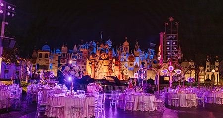An evening reception setup with round tables and chairs in front of the illuminated 'it's a small world' facade