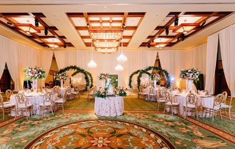An elegant banquet room adorned with floral centerpieces and archways, featuring round tables set for guests under chandeliers and draped curtains
