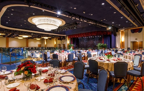 An indoor banquet hall features round tables and floral centerpieces organized in front of a stage with dramatic lighting and a chandelier