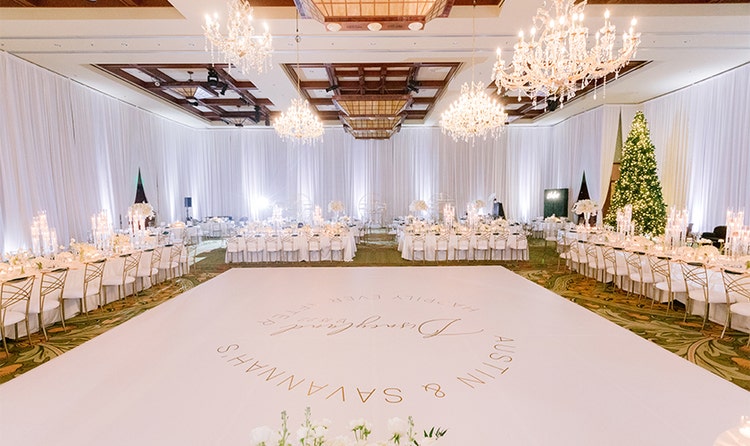 A spacious dance floor in the center of a ballroom featuring high ceilings, long curtains and chandeliers