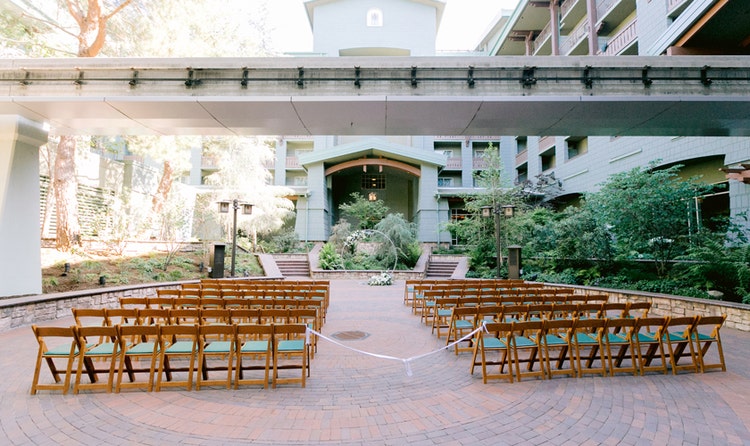 Rows of outdoor seating on a cobblestone courtyard leading to a wedding arbor beneath a monorail