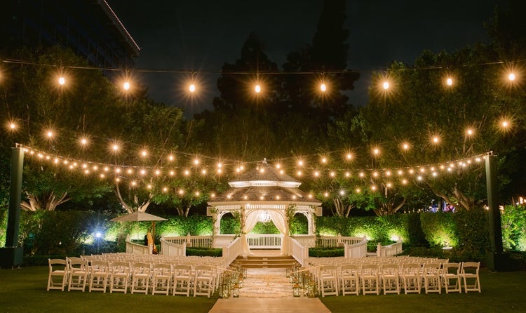 An outdoor wedding venue at night, featuring a pergola and rows of folding chairs beneath strings of lights A pergola in an open garden setting strung with lights