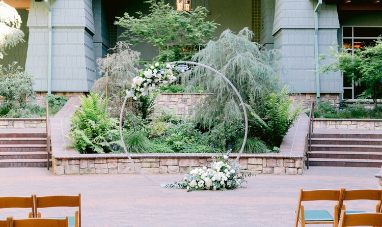 A round wedding arbor adorned with 2 floral arrangements in front of foliage with a hanging lantern