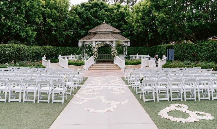 At an outdoor wedding venue, rows of folding chairs facing a pergola flank a carpeted aisle accented with flower petals arranged in decorative shapes