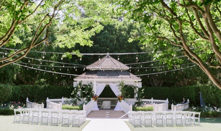 In a garden setting, rows of folding chairs face a pergola set for a wedding, with fabric draping pulled to the side to frame the stage