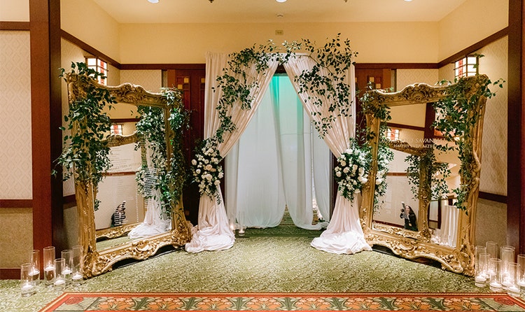 A curtain covered with ivy and flowers with 2 standing mirrors on either side surrounded by candles