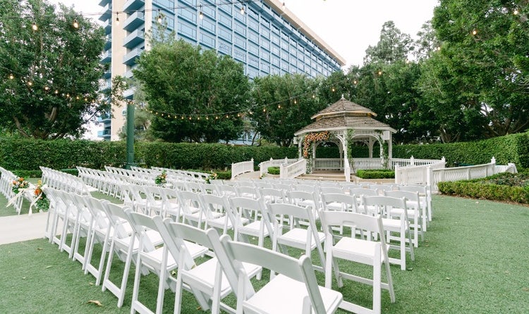 At an outdoor wedding venue, rows of folding chairs facing a pergola are separated by an aisle