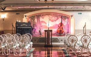 A cello, saxophone and piano on a stage before rows of chairs flanking an asle