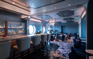 An elegant lounge with bar seating, porthole windows, a crystal chandelier and a grand piano at Nightingale's aboard the Disney Wish