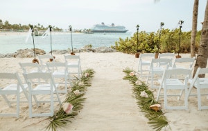 A beachfront wedding setup featuring 2 rows of chairs and an aisle decorated with palm leaves and shells