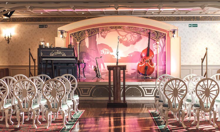 A room with rows of chairs set in front of a podium features a stage with a piano and jazz instruments on stands