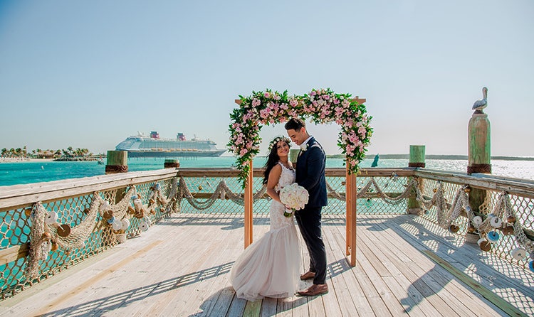 A bride and groom stand under a flower covered archway on a dock