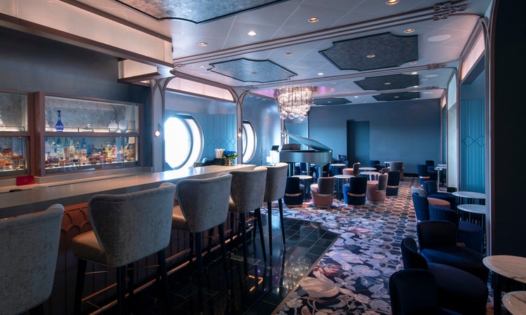 An elegant lounge with bar seating, porthole windows, a crystal chandelier and a grand piano at Nightingale’s aboard the Disney Wish