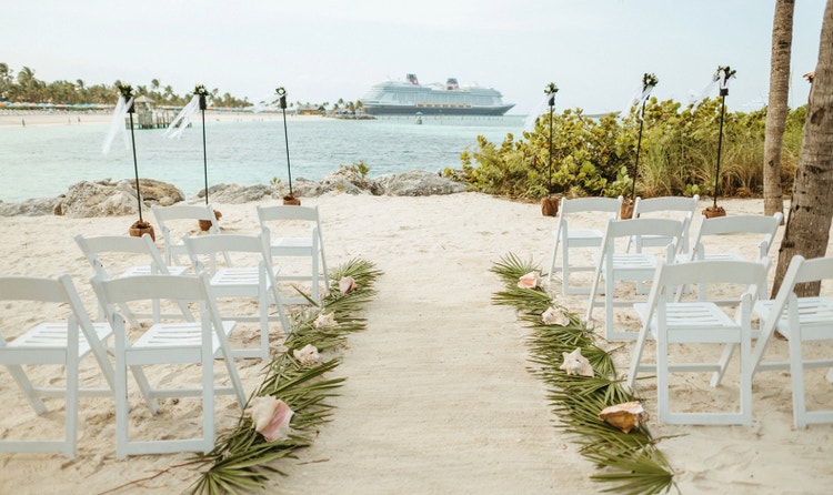 Beachside wedding ceremony setup with palm fronds topped by shells lining the sandy aisle and a Disney cruise ship nearby