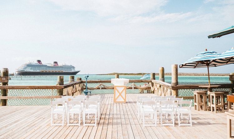 Wedding ceremony site on a wooden pier with chairs forming an aisle, sun umbrellas off to the side and a cruise ship in view