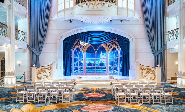 A lavish atrium set for a wedding ceremony aboard a Disney cruise ship with rows of chairs and floral arrangements flanking an aisle