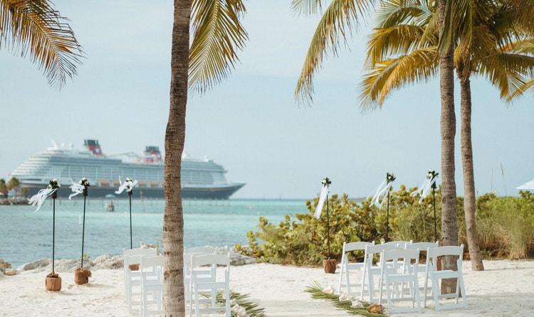 Intimate beach wedding setup framed by palm trees with ceremony seating, a sandy aisle with greenery and a cruise ship at sea