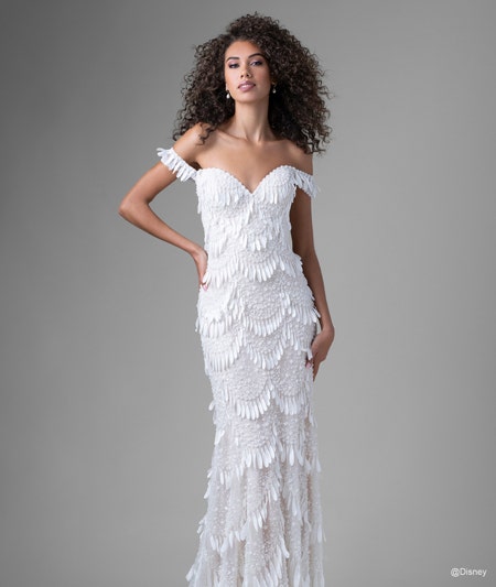 The front of a bride wearing a Tiana inspired sheath gown featuring an off the shoulder neckline and light fringe detail