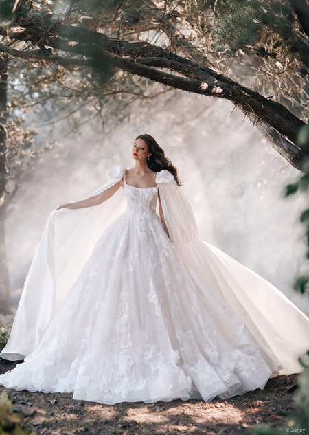 A woman posing in a forest wearing a fairy tale wedding gown with floral details and a flowing cape with puffed sleeves