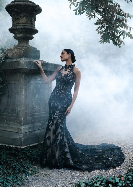 A woman posing by a stone structure in the forest wearing a fitted gown with lace details and a long train