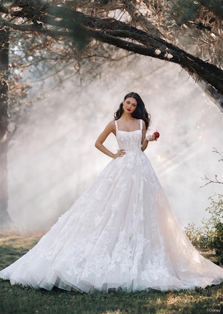 A woman posing in a forest wearing a fairy tale wedding gown with floral details holding an apple