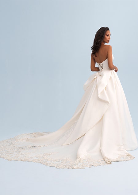 A woman looking over her shoulder wearing a wedding dress with a long and lacy trail and detachable bow on the back