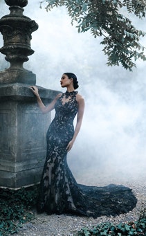 A woman posing by a stone structure in the forest wearing a fitted gown with lace details and a long train