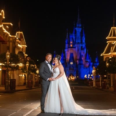 At night, on Main Street U S A, a couple in wedding attire holds hands, with Cinderella Castle illuminated in the background
