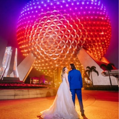 A couple in wedding attire holds hands in front of the illuminated Spaceship Earth geosphere at Epcot, at night