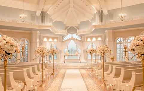 Sunlight shines through the arched stained glass windows of a wedding chapel with chandeliers and a high beam ceiling