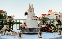 A two tiered wedding cake with a Cinderella Castle cake topper on a table adorned with flowers and silver tea lights