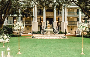 A stately manor with oak trees and a fountain is set up for an outdoor wedding on the lawn