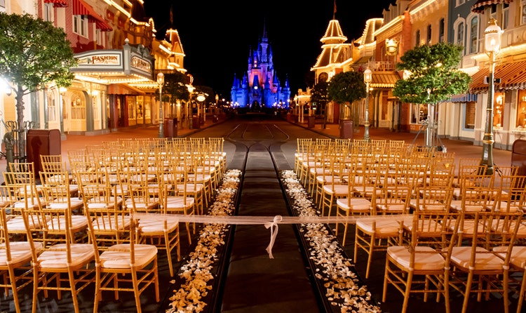 An evening wedding ceremony venue with bamboo chairs and a flower petal lined aisle, set on Main Street USA with Cinderella Castle in the background