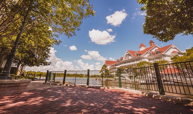 A shady brick path with a cast iron guardrail and old fashioned streetlamps looks out to the Seven Seas Lagoon and Disney's Grand Floridian Resort & Spa