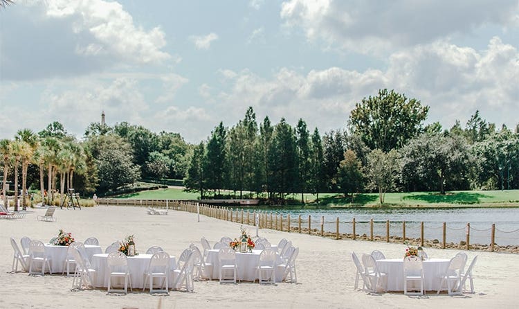 Four tables surrounded by chairs on a beach with trees in the background