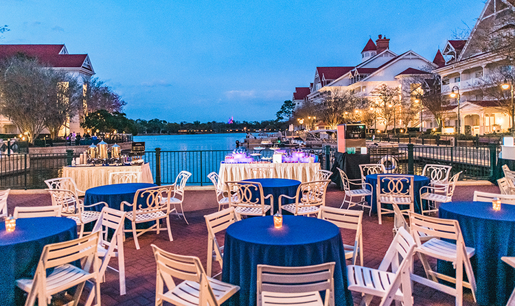 The patio at Disney’s Grand Floridian Resort and Spa, overlooking the marina at dusk, with many small tables covered with tablecloths, and folding chairs around the tables