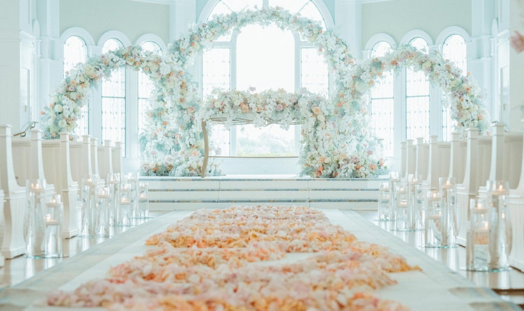 Three floral arches on an altar at the end of an aisle runner with rose petals near 2 rows of pews and vases with floating candles