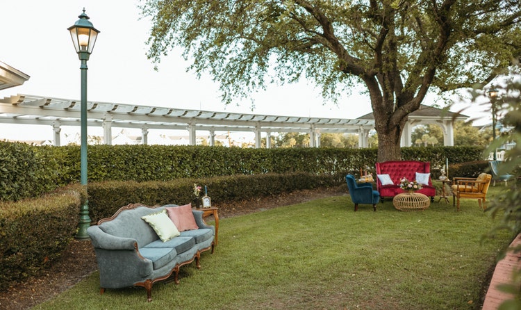 Velvet sofas and armchairs on a lawn beside a hedge and a tree at a wedding cocktail hour at Seabreeze Point