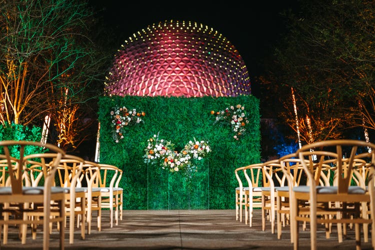 Rows of wooden chairs and a garden wall with 3 floral arrangements in front of Spaceship Earth