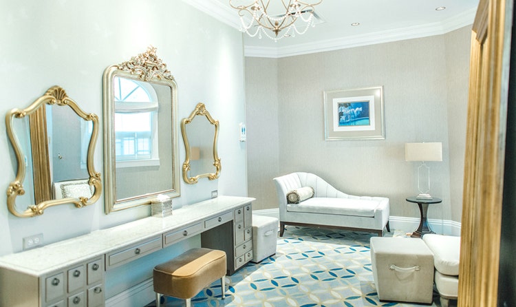 An elegant bridal suite with a vanity dresser, 3 mirrors, a cushioned bench, a chandelier and a chaise longue