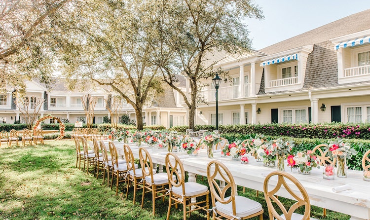 A garden venue decorated for a wedding reception with a long table, chairs and flowers in vases at Poolside Green at Disney’s BoardWalk Inn