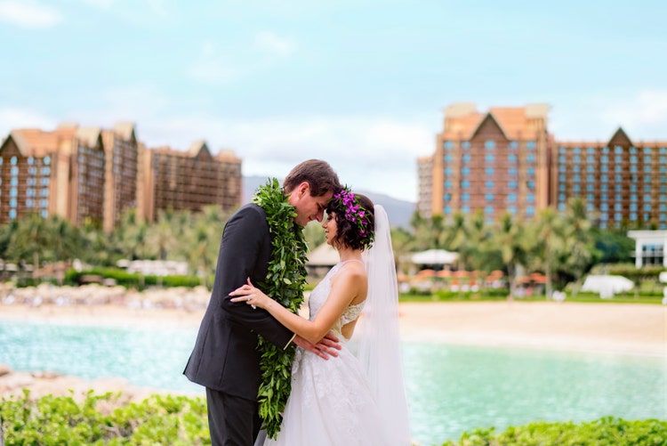 A bride wearing a floral headpiece, smiling at a groom wearing a maile lei