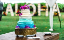A 3 tiered wedding cake with piped frosting and tropical flowers on a table in front of the words, ‘Aloha’