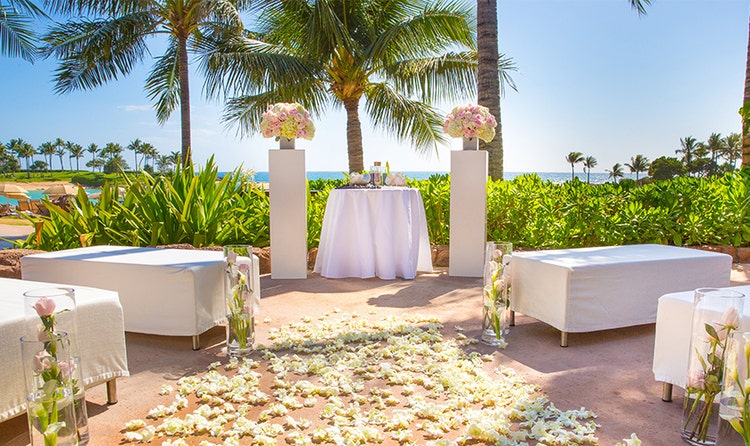 In a beachfront garden at Aulani Resort, banquettes and glass vases are arranged in front of an altar that is flanked by pillars topped with rose bouquets