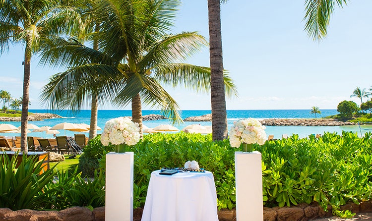 By the beach at Aulani Resort the wedding altar of Sunset Alcove features a table holding a book, 2 kukui nut leis and a conch shell with a pillar supporting a rose bouquet to either side