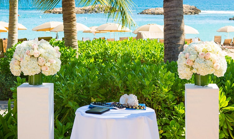 In a garden setting, 2 pillars topped with rose bouquets flank a table holding a book, 2 kukui nut leis and a conch shell
