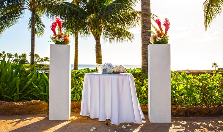 With the beach at Aulani Resort in the background, the wedding altar of Sunset Alcove features a table holding 2 conch shells with a pillar supporting a flower arrangement to either side