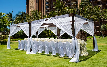 A canopy covers a long table in the middle of a lawn near a hotel building