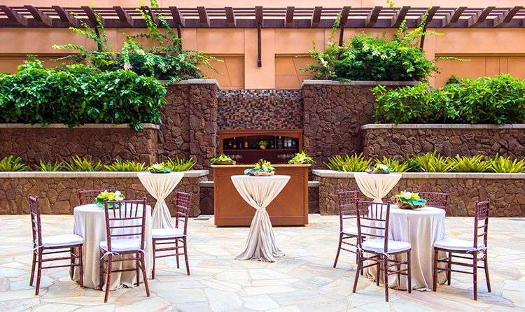 Dining tables and cocktail tables decorated for a wedding in a resort courtyard, against a rustic, stone-wall backdrop