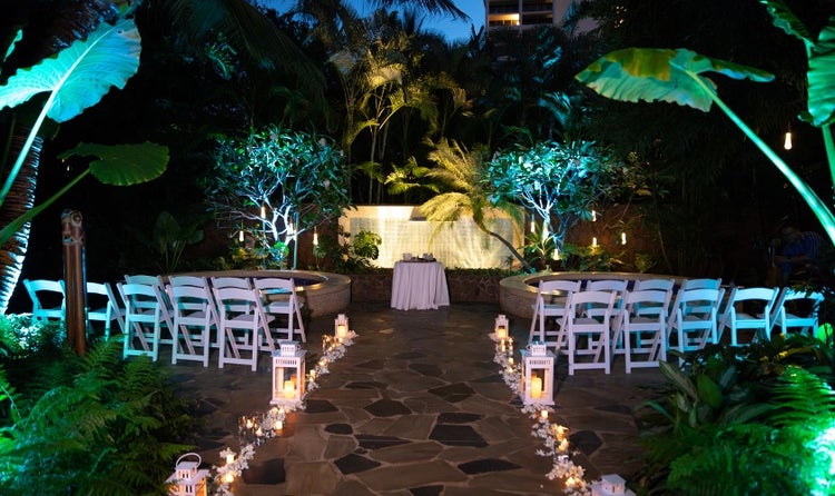 A walkway of flowers and candles leads past chairs toward a table in front of a fountain